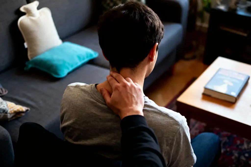 Une personne assise se masse la nuque dans un salon confortable, livre sur la table.