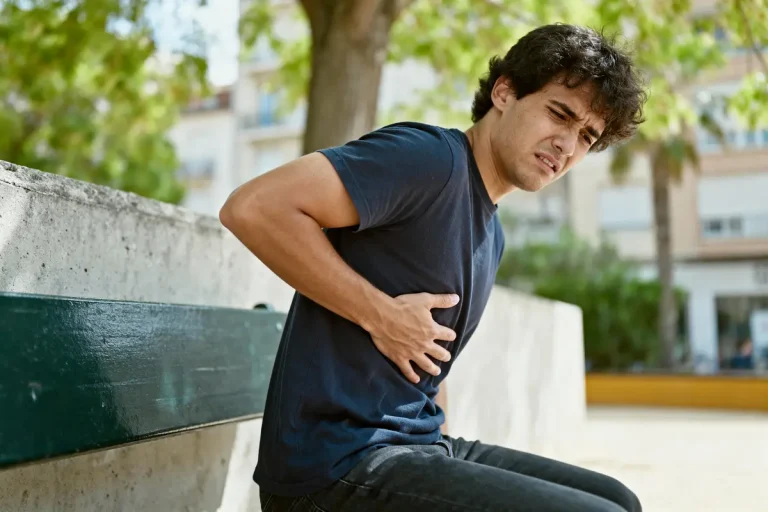 Homme en t-shirt bleu grimaçant, tenant son côté, assis sur un banc en plein air, arbres en arrière-plan.