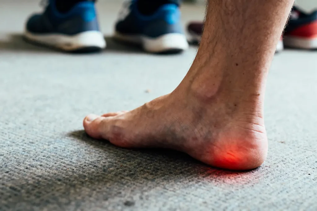 Talons nus sur tapis gris, douleur au talon éclairé par une lumière rouge. Chaussures floues à l’arrière.