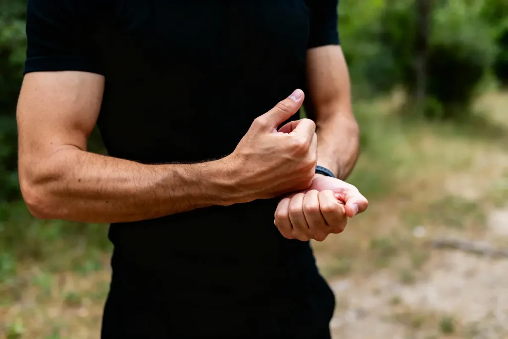Homme en noir, extérieur, tient son poignet gauche avec sa main droite. Arrière-plan flou de verdure.