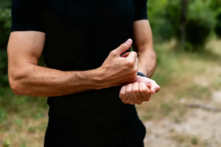 Homme en noir, extérieur, tient son poignet gauche avec sa main droite. Arrière-plan flou de verdure.