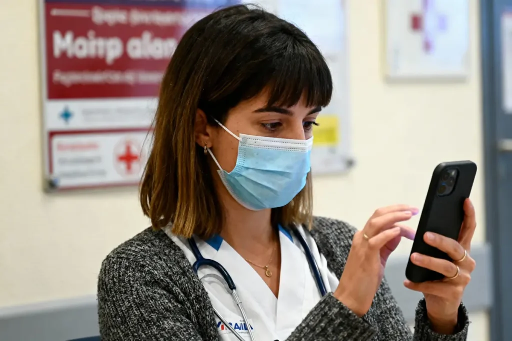 Femme masquée en blouse médicale utilise un smartphone, stéthoscope autour du cou, dans un environnement hospitalier.