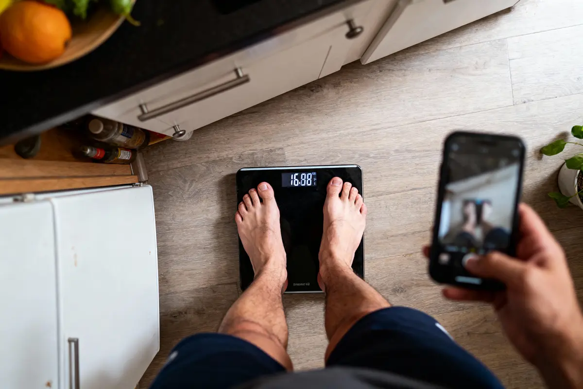 Personne debout sur une balance, prenant une photo avec un smartphone dans une cuisine, affichage lumineux.