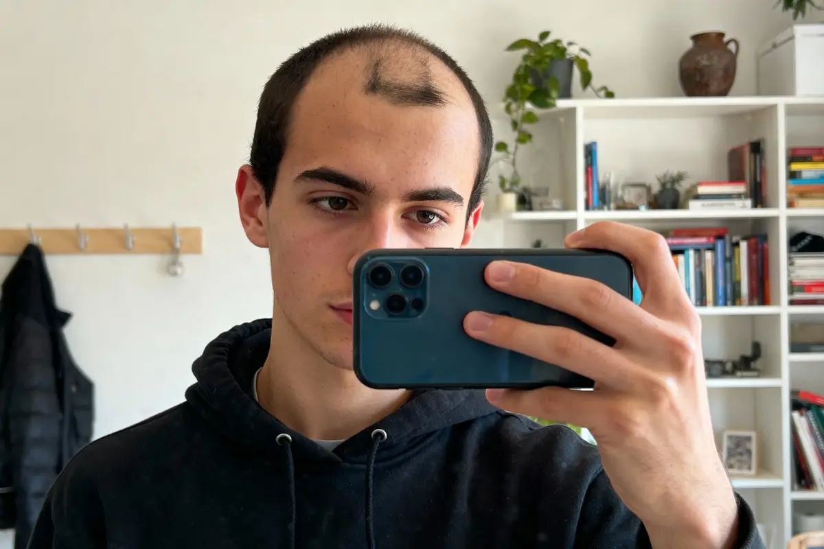 Jeune homme prenant un selfie, cheveux clairsemés, portant un sweat à capuche noir, intérieur avec étagères et livres.