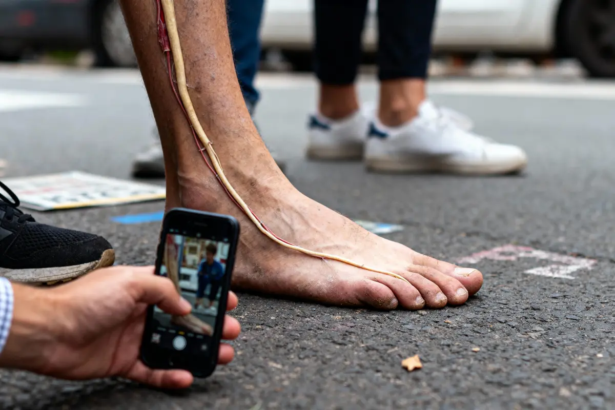 Pied nu avec câbles, photographié par une personne tenant un smartphone sur une rue pavée.