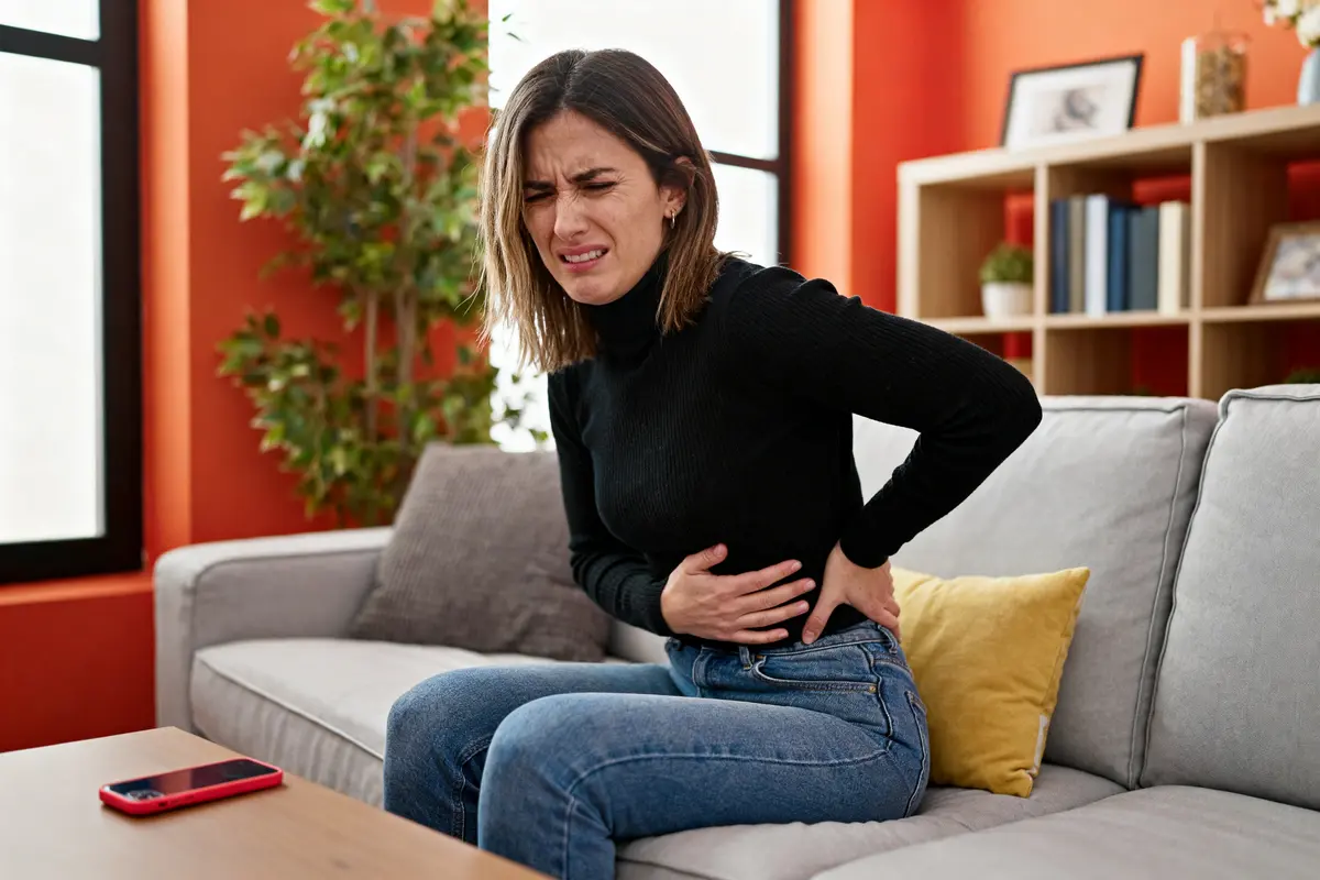 Femme assise sur un canapé, souffrant de douleurs abdominales, main sur le ventre, ambiance intérieure lumineuse.