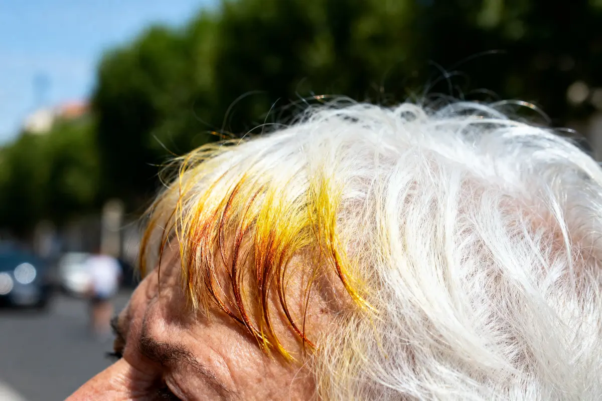 Cheveux blancs avec mèches jaunes et oranges, en extérieur ensoleillé, arrière-plan flou avec arbres.