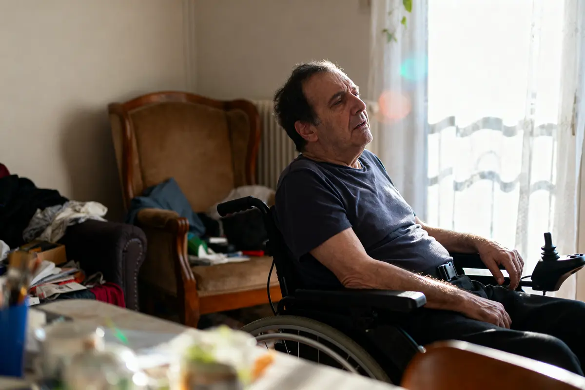 Homme en fauteuil roulant près de la fenêtre, éclairé par la lumière du jour, dans une pièce désordonnée.